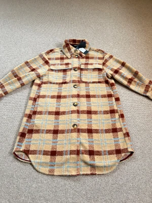 Soaked in Luxury – Whey & Mahogany Checkered Coat (NY med tags) Kappa - Lyxig och stilsäker rutmönstrad kappa från Soaked in Luxury, i färgerna Whey (kräm/beige) och Mahogany (djup brun). Den klassiska rutdesignen kombinerar moderna toner med tidlös elegans – en perfekt kappa för både vardag och mer uppklädda tillfällen.  Kappan har en rak, strukturerad silhuett som ger ett sofistikerat uttryck. Materialet är mjukt men ändå stadigt, vilket ger ett fint fall och gör den skön att bära. De subtilt markerade axlarna, de diskreta knapparna och det välarbetade mönstret. 