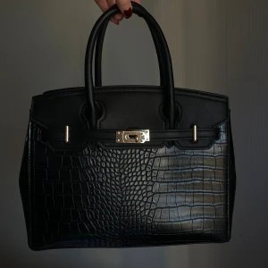 Svart handväska med krokodilmönster - Stilren svart handväska med krokodilmönstrad yta och snygga guldfärgade detaljer. Väskan har två rejäla handtag, en dekorativ låsdetalj framtill och en praktisk dragkedja på baksidan. Perfekt storlek för att få plats med allt du behöver. (Medel size)