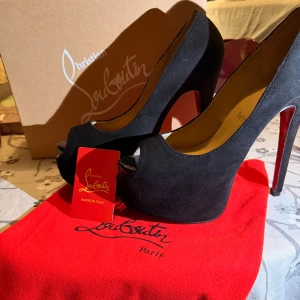 Christian Louboutin svarta pumps - Svarta pumps från Christian Louboutin med ikonisk röd sula, hög klack och öppen tå. Skorna är tillverkade i mocka och har en elegant plattform framtill. Perfekt för dig som vill sticka ut med en lyxig och klassisk look.