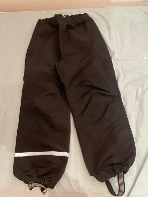 Svarta täckbyxor med reflexdetalj - Svarta täckbyxor med elastisk midja och reflexdetalj på ena benet. Byxorna har resår och justerbara fothällor vid bensluten för extra komfort och skydd mot regn snö och kyla. Perfekta för kalla dagar utomhus.