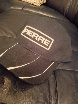 Svart keps från Pierre med vita detaljer - Svart keps från Pierre med broderad logga framtill i vitt. Skärmen har coola vita linjer som ger en sportig vibe. Klassisk form med böjd skärm och ventilerande hål upptill. Perfekt för dig som gillar enkel och stilren streetwear.