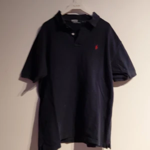 Matinblå Polo Ralph Lauren piké XL - Klassisk marinblå pikétröja från Polo Ralph Lauren i storlek XL. Tröjan har korta ärmar, krage och knappar framtill. På bröstet finns den ikoniska röda broderade Polo-loggan. Materialet är mjuk bomull som känns skön mot huden.