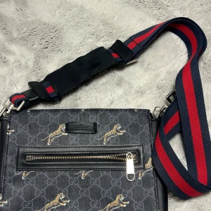 Gucci axelväska med tigerprint - Snygg axelväska från Gucci med mörkgrå bakgrund och klassiskt GG-mönster, dekorerad med tigrar. Väskan har en bred axelrem i marinblått och rött, samt en praktisk dragkedja framtill. Materialet är canvas med detaljer i skinn.