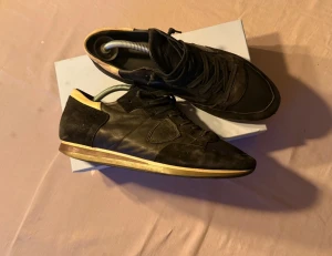 Svarta sneakers från Philippe Model - Säljer ett par svarta sneakers från Philippe Model med snygga detaljer i skinn och mocka. Skorna har snörning, rund tå och en beige kant på sulan som ger en cool kontrast. Perfekta för dig som gillar stilrena och trendiga sneakers.