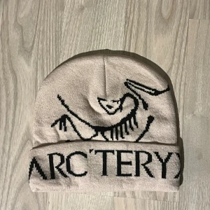 Beige Arcteryx mössa med logga - Snygg beige mössa från Arcteryx med svart logotyp och coolt fossilmotiv framtill. Perfekt för dig som gillar outdoor och vill ha en stilren look. Mjuk och skön passform som håller dig varm under kyliga dagar.