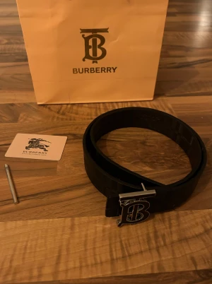 Svart Burberry bälte - Säljer ett svart Burberry bälte i skinn med klassiskt rutigt mönster och präglad riddare. 
