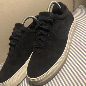 Svarta mocka sneakers från Axel Arigato - Snygga svarta sneakers från Axel Arigato i mjuk mocka med vit platt sula. Klassisk låg modell med rund tå och svart snörning. Perfekt för dig som gillar stilrena och minimalistiska skor med premiumkänsla.