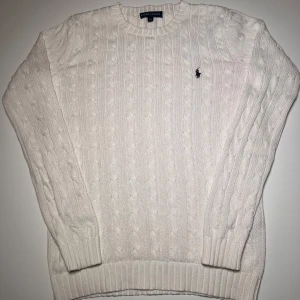 Vit kabelstickad tröja Ralph Lauren - Snygg vit kabelstickad tröja från Ralph Lauren med klassisk rund hals och broderad logga på bröstet. Tröjan har långa ärmar och ribbade muddar vid hals, ärmslut och nederkant. Perfekt för dig som gillar stilrena och tidlösa plagg.