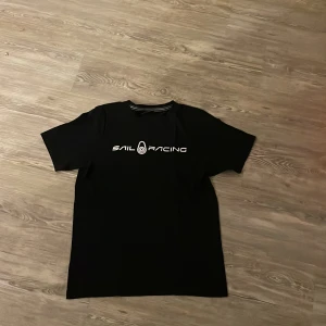 Svart Sail Racing t-shirt med tryck - Svart t-shirt från Sail Racing med vitt logotryck på bröstet och en mindre logga i nacken. Klassisk rund hals och korta ärmar. Materialet är mjuk bomull som känns skön mot huden. Perfekt för dig som gillar stilrena och sportiga plagg. Skriv vid minsta fundering ✅