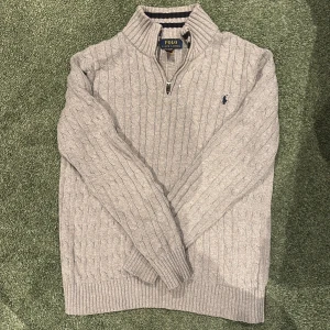 Grå stickad tröja Polo Ralph Lauren - Snygg grå stickad tröja från Polo Ralph Lauren i storlek 170 cm. Tröjan har kabelstickat mönster, ribbad krage och muddar samt en half zip-dragkedja framtill. Klassisk broderad logga på bröstet och långärmad modell. Perfekt för dig som gillar stilren och tidlös design. Pris kan diskuteras