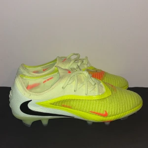 Nike Phantom 6 elite voltage fotbollsskor  - Nike Phantom fotbollsskor i en fräsch mix av ljusgul, vit och neonorange. Skorna har snörning, stickad ovandel och tydliga Swoosh-loggor i svart och orange. Perfekta för dig som vill sticka ut på planen med både stil och funktion.