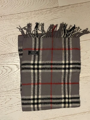 Grå rutig Burberry halsduk i kashmir - Snygg halsduk från Burberry i mjuk kashmir. Klassisk rutig design i grått med svarta, vita och röda linjer. Lång och smal modell med fransar i ändarna, perfekt för att ge din outfit en lyxig touch.