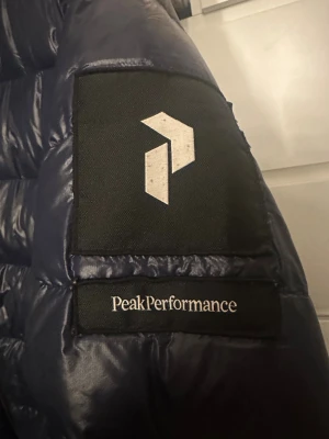 Mörkblå Peak Performance dunjacka - Snygg mörkblå dunjacka från Peak Performance med huva och dragkedja framtill. Jackan har quiltad design och logotyp på ärmen. Perfekt för kalla dagar och har en modern, sportig look. Fickor med dragkedja på sidorna och justerbar huva.