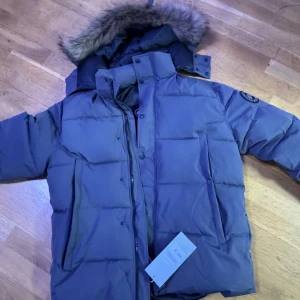Grå dunjacka från Canada Goose S - Säljer en grå dunjacka från Canada Goose med fet pälskantad huva och klassisk patch på ärmen. Jackan har dragkedja, knappar och stora fickor framtill. Perfekt för kalla vinterdagar och riktigt snygg streetstil.