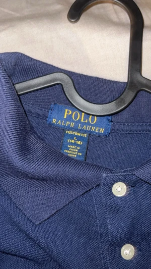 Marinblå pikétröja från Ralph Lauren - Snygg marinblå långärmad pikétröja från Ralph Lauren med klassisk krage och tre vita knappar framtill. Tröjan har det ikoniska röda Polo-logot broderat på bröstet och är gjord i mjuk bomull. Perfekt för dig som gillar stilrena och tidlösa plagg.