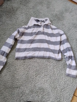 Randig croppad pikétröja Gina Tricot - Snygg croppad pikétröja från Gina Tricot i storlek XS. Tröjan har breda grå och vita ränder, klassisk krage och två knappar framtill. Sparsamt använd.