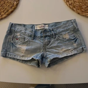 Ljusblå jeansshorts från Hollister - Säljer ett par ljusblå lågmidjade jeansshorts från Hollister med slitna detaljer och råa kanter. Klassisk femficksmodell med låg midja och knappgylf. Perfekta för varmare dagar. 