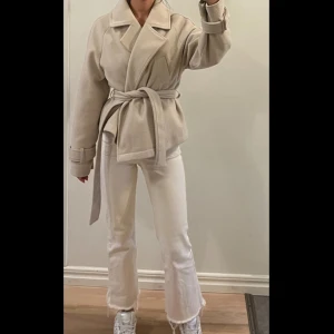 Beige omlott kappa från Nelly - Snygg beige kappa från Nelly med bred krage och omlottmodell. Jackan har ett bälte i midjan och dekorativa spännen vid ärmsluten. Tillverkad i mjukt ullmaterial som ger en lyxig känsla. Perfekt för dig som gillar stilrena och trendiga plagg. Jackan är lite nopprig