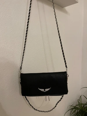 Svart axelväska zadig voltaire - Snygg svart axelväska i skinnimitation med silverfärgad kedja och detaljer. Framsidan har ett emblem med glittrande vingar och två dragkedjor. Baksidan har en extra ficka med dragkedja. Perfekt storlek för mobilen och det viktigaste.