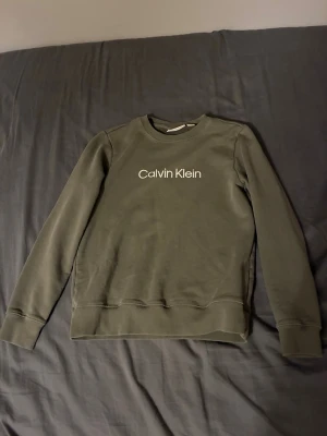 Olivgrön Calvin Klein sweatshirt - Snygg olivgrön sweatshirt från Calvin Klein med vit logotyp broderad på bröstet. Tröjan har rund hals, ribbade muddar och lång ärm. Perfekt för dig som gillar stilrena och klassiska plagg med en modern touch.