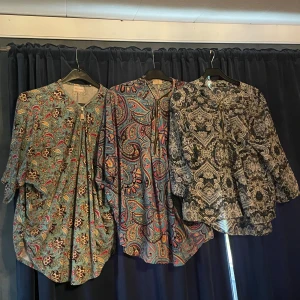 Tre färgglada henley-skjortor med mönster - Tre henley-skjortor med unika mönster: en blågrön med blommor och paisley, en färgstark paisley i blått, rött och gult, samt en svartvit skjorta med detaljerat paisley-mönster. Alla har knappar vid halsen och lång ärm. Perfekt för dig som gillar boho-vibe och coola prints.