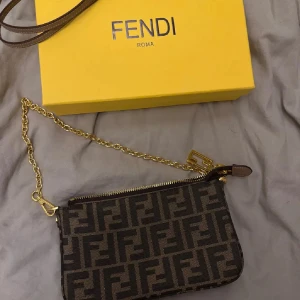 Fendi axelväska med FF-mönster - Snygg axelväska från Fendi med ikoniskt FF-mönster i mörkbrunt och svart. Väskan har guldfärgad kedja, dragkedja och detaljer i skinn. Perfekt storlek för det viktigaste och har en innerficka. Kommer med originalkartong och tag.