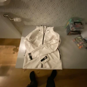 Vit vindjacka från Moncler med huva - Snygg vit vindjacka från Moncler med svart dragkedja och justerbar huva. Jackan har Moncler-logga på ärmen och praktiska detaljer som kardborre vid ärmslut. Perfekt för dig som gillar clean och sportig stil.