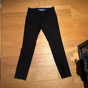 Svarta kostymbyxor  - Snygga svarta kostymbyxor med smal och rak siluett. Klassisk design med bälteshällor, sidfickor fram och två bakfickor med knapp. Perfekta för en stilren look och passar till skjorta eller t-shirt. Materialet känns mjukt och har en lätt stretch.