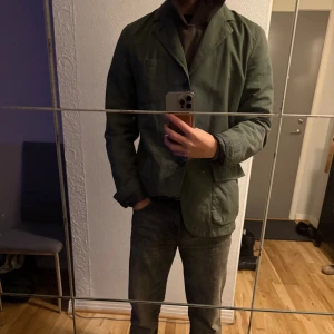 Woolrich Kavaj - Snygg grön kavaj med klassisk krage och två stora fickor framtill. Jackan har knäppning med knappar och är i en rak, normal passform. Materialet känns som bomull och den har långärmade ärmar. Perfekt för dig som gillar stilren och enkel design. Storlek M passar L