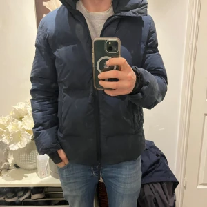 Mörkblå pufferjacka från A Day's March - Säljer en mörkblå pufferjacka från A Day's March med huva och dragkedja. Passar perfekt nu till vintern. Ända anmärkningen jag har är höger ärms ”handvärmare” där sömmarna har släppt. Något som enkelt kan fixas med nål och tråd. Det är dock inget som påverkar varken jackans funktion eller utseende.
