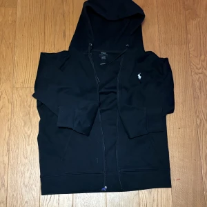 Svart zip från Polo Ralph Lauren - Svart hoodie från Polo Ralph Lauren med klassisk broderad logga på bröstet. Tröjan har huva med snörning, dragkedja framtill och två fickor. Materialet är mjuk bomull och passformen är normal. Perfekt för chill dagar och streetwear. Säljer pågrund av att jag int använder den längre rensar min garderob! 