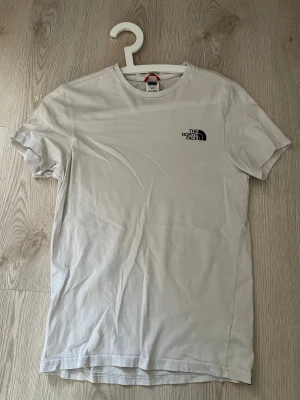 Vit t-shirt från The North Face XS - Snygg vit t-shirt från The North Face i storlek XS. Klassisk rund hals och korta ärmar, med svart logga tryckt på bröstet. Tillverkad i mjuk bomull som känns skön mot huden. Perfekt för dig som gillar stilrena och enkla plagg.