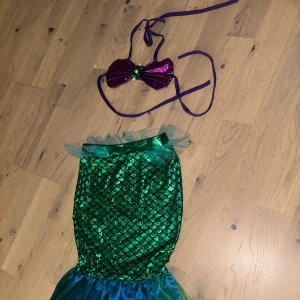 Grön sjöjungfruklänning 3 år Sjöjungfru-kostym - Superfin sjöjungfruklänning för barn, med glittrig grön kjol i fiskfjällsmönster och blågrön volang nertill. Toppen är lila med snörning och pärldetaljer. Perfekt för att känna sig som en riktig havsprinsessa!Fint sjöjungfru-set med en kjol i grönt och blått med detaljer i tyll. Överdelen är lila.