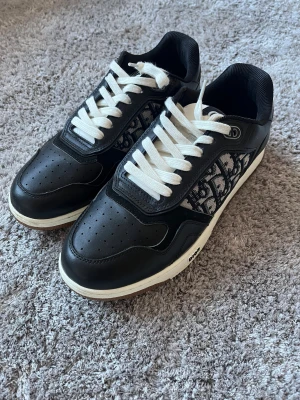  Dior b27 - Snygga svarta Dior sneakers med vit sula och vita snören. Sidorna har det klassiska Dior-monogrammet i svart och vitt. Skorna är i skinn och textil, med perforerad tå och rund form. Perfekta för dig som vill ha en lyxig och trendig look.