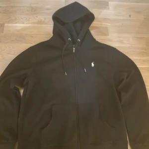 Svart hoodie från Polo Ralph Lauren - Svart hoodie från Polo Ralph Lauren med dragkedja framtill, snörning i huvan och klassisk vit logga på bröstet. Tröjan har kängurufickor och är tillverkad i mjukt bomullsmaterial. Perfekt för en chill och stilren look. HELT NY!