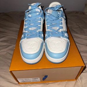 Louis Vuitton blå  - Säljer ett par Louis Vuitton sneakers i ljusblått och vitt med klassisk LV-logga på tån och sidan. Skorna har snörning, platt sula och rund tå. Tillverkade i skinn och textil, med detaljerade mönster på sidorna och sulan. Riktigt snygga och unika sneakers för dig som gillar exklusiva märken.