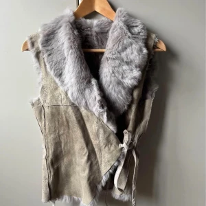 Grå pälsväst med omlott och knyt - Säljer en snygg grå pälsväst med omlott-design och knytband i sidan. Västen är tillverkad i mjuk äkta päls med mockaliknande utsida. Går att använda med antingen päls eller skinnsidan utåt. 