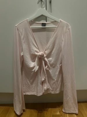 Ljusrosa omlottopp med knyt från Gina Tricot - Supersöt ljusrosa omlottopp från Gina Tricot med lång ärm och v-ringning. Toppen har ett snyggt knyt framtill som ger en chill och feminin vibe. Mjuk och skön bomullskvalitet, perfekt för att lyfta din outfit med lite extra stil. (Liten i storleken!! Passar som M)