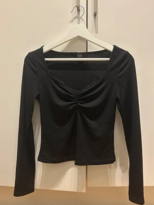 Svart långärmad topp  - Svart långärmad topp från med snygg v-ringning och rynkad snörning framtill. Toppen har ribbad struktur och spetsdetaljer vid halsen som ger en extra touch. Perfekt för dig som gillar stilrena och trendiga plagg.