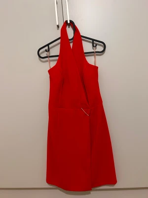 Röd halterneck klänning från Calore - Snygg röd kortklänning från Calore med halterneck och omlott-detalj framtill. Klänningen har en slät yta med glansig insida och dragkedja i ryggen. Passformen är normal och materialet känns som polyester, perfekt för dig som vill sticka ut med en färgstark look.