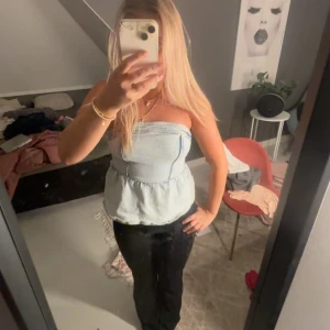 Ljusblå axelbandslös tubtopp - Säljer en ljusblå axelbandslös tubtopp i denim-look med volangkant nedtill. Toppen har bandeau-modell och markerad söm i midjan för en snygg passform. 