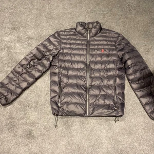 Svart dunjacka från Ralph Lauren - Säljer en svart dunjacka från Ralph Lauren med diskret röd logga på bröstet. Jackan har dragkedja framtill, två sidofickor med dragkedja och justerbar dragsko nedtill. Perfekt för kyliga dagar och riktigt lätt att bära.