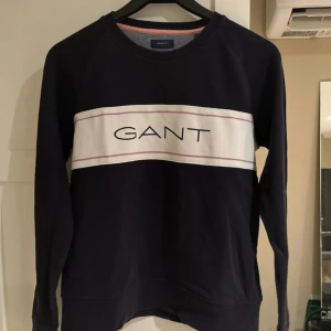 Mörkblå GANT sweatshirt med logga - Aldrig använd! Snygg mörkblå sweatshirt från GANT med vit panel och rosa linjer över bröstet samt stor GANT-logga. Tröjan har rund halsringning och långa ärmar. Tillverkad i mjuk bomull, perfekt för en chill och stilren look. Djur och rökfritt hem! 