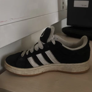 Adidas Campus svarta sneakers - Svarta Adidas Campus sneakers med klassiska vita ränder på sidorna och vit Adidas-logga på hälen och plösen. Skorna har snörning och en platt sula i gummi. Ovandelen är i mocka och insidan är ljusgrå. Perfekt för dig som gillar retrostil och streetwear.