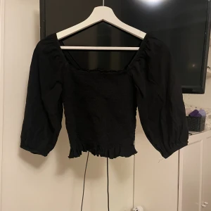 Svart smockad blus från Lindex - Supersöt svart blus från Lindex i smockat tyg med fyrkantig ringning och trekvartsärmar. Blusen är croppad med volangkant nertill och har en lätt puffig ärm för en trendig look. Perfekt att styla med både jeans och kjol.