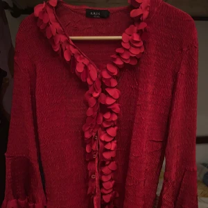 Röd kofta med volangdetaljer från Kris - Unik röd kofta från Kris med coola volangdetaljer och dekorativa applikationer längs krage och ärmslut. Koftan har en lätt skrynklig struktur och knäppning framtill. Perfekt för dig som vill sticka ut med en färgstark och trendig look.