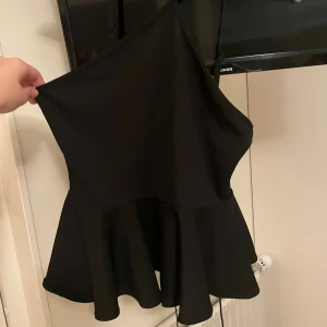 Svart peplumtopp från NA-KD - Supersnygg svart peplumtopp från NA-KD med asymmetrisk halsringning och smala axelband som korsas framtill. Toppen har en volang nertill som ger en cool siluett. Aldrig använd och lappen är kvar 