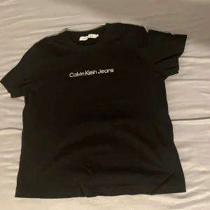 Svart Calvin Klein Jeans t-shirt - Svart t-shirt från Calvin Klein Jeans med vit logga tryckt på bröstet. Klassisk rund halsringning och korta ärmar. Tillverkad i mjuk bomull för en skön och avslappnad känsla. Perfekt för en clean och stilren look.
