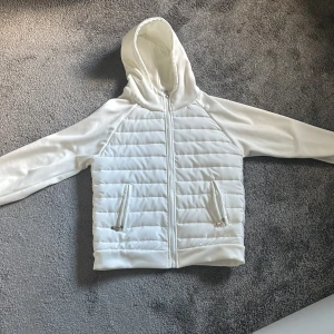 Vit cardigan - Säljer en stilren vit pufferjacka med huva och dragkedja framtill. Jackan har quiltade partier på kroppen och släta ärmar, samt två fickor med dragkedja. Den är aldrig använd och har inga skador, pris kan diskuteras vid snabb affär.