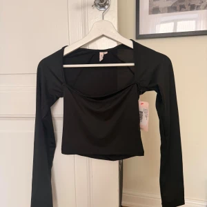 Svart långärmad topp från Nelly  - Svart långärmad topp från Nelly i storlek XS. Toppen har en snygg croppad passform med fyrkantig urringning och är tillverkad i ett stretchigt, slätt material. Perfekt att styla med jeans eller kjol för en trendig look.